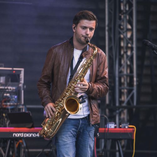 Ein Mann spielt konzentriert ein Saxophon auf einer Bühne. Er trägt eine braune Lederjacke, ein weißes T-Shirt und Jeans. Im Hintergrund sind Musikequipment und ein rotes Keyboard (Nord Piano 4) zu sehen. Die Szene wirkt ruhig und fokussiert, mit professionellem Bühnenlicht.