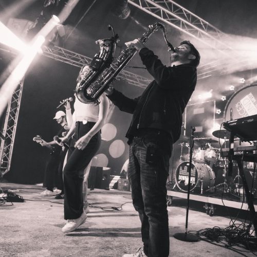 Eine Live-Band performt auf einer Bühne in stimmungsvoller Atmosphäre. Im Vordergrund spielt ein Musiker leidenschaftlich Saxophon, daneben tanzt eine Sängerin mit Mikrofon in der Hand. Weitere Bandmitglieder sind mit Gitarre und Schlagzeug im Hintergrund zu sehen. Die Szene ist in Schwarz-Weiß gehalten und von Scheinwerfern beleuchtet.